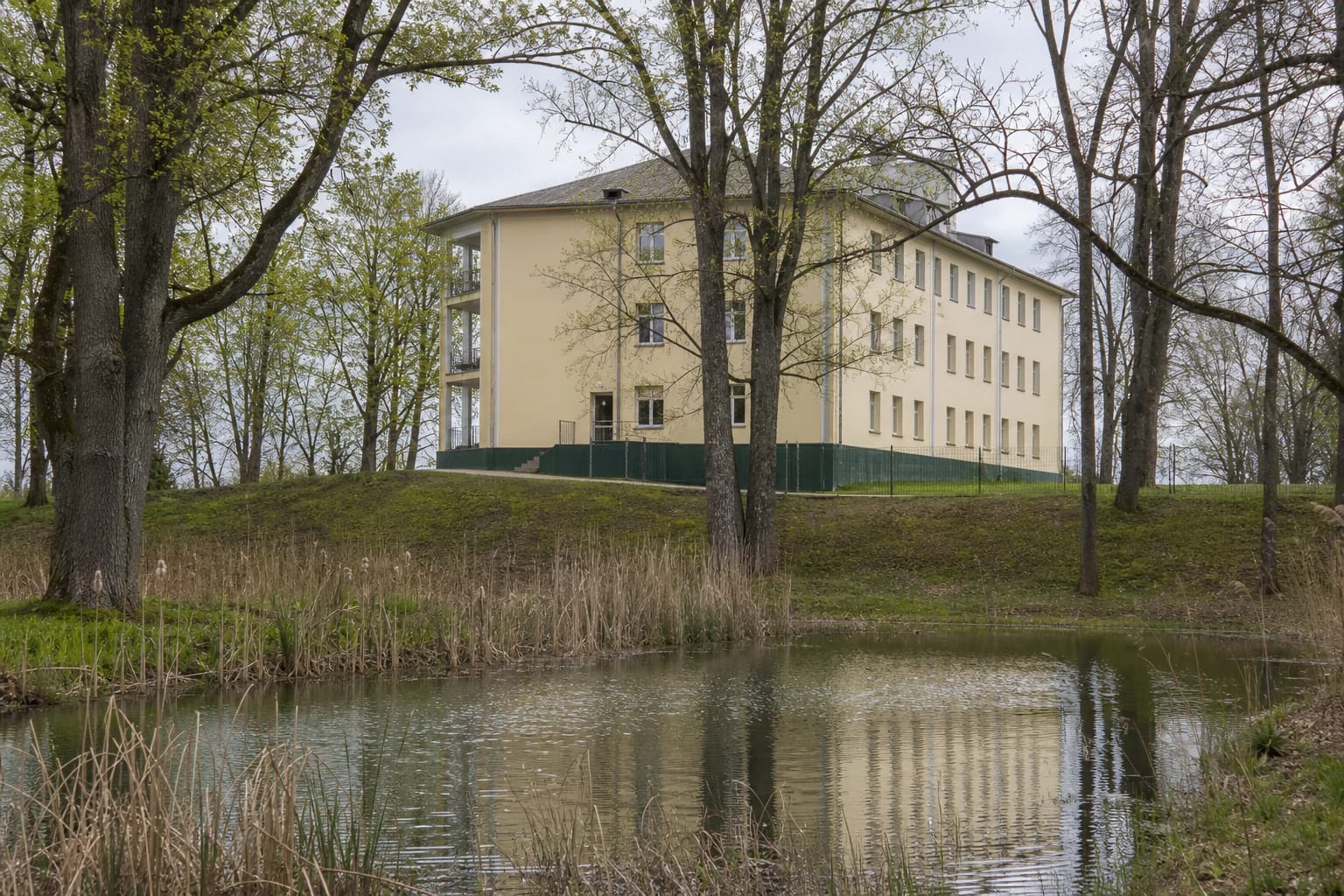 Light-coloured brick Adakavas Manor palace beyond a pond and old park trees
