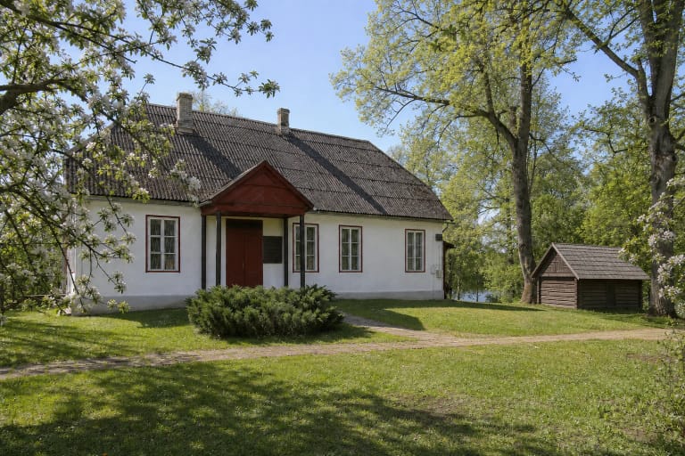 Белый дом-музей Адомаса Хребницкиса с красным крыльцом, отдельный деревянный домик-клеть и цветущие яблони в зелёном парке Роюса