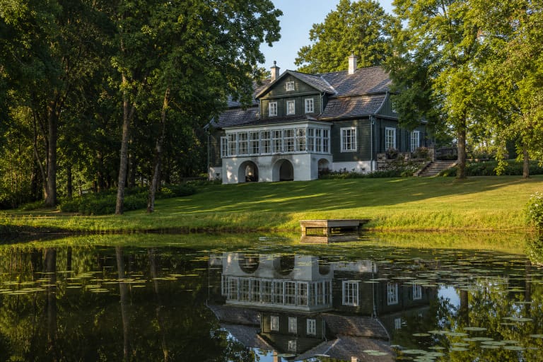 Wooden Adomynė Manor with white arched foundations reflected in a park pond