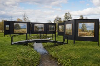 AJJU Open-Air Gallery paintings on dark panels above a stream and black arched bridge