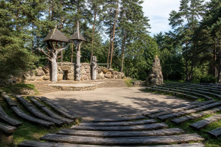 Stone outdoor amphitheatre in Akmenė Nature and Culture Park with boulder walls, rustic timber towers, and benches beneath tall pines