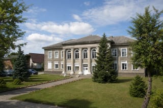 Grey two-storey Akmenė Regional Museum with pale pilasters, arched windows, and a green square in front
