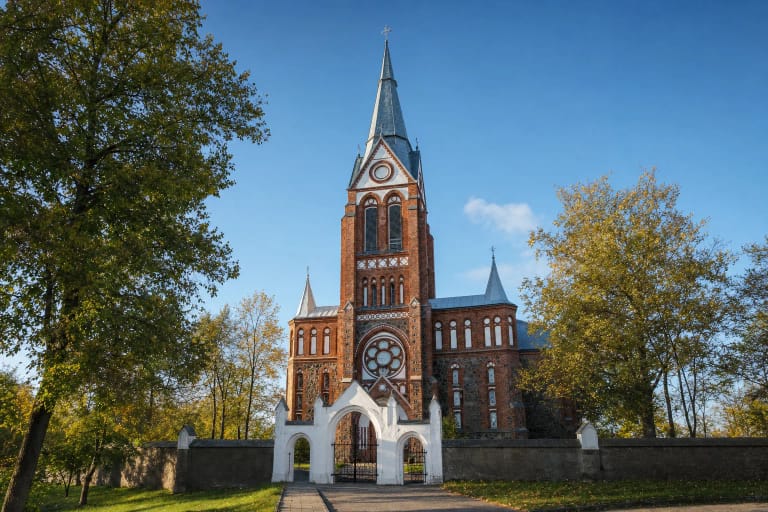 Akmenės Šv. Onos bažnyčios aukštas raudonų plytų bokštas, lauko akmens sienos, balti vartai ir apskritas langas rožė rudens šviesoje
