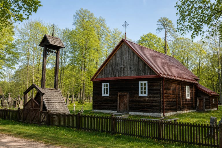 Dark-log Akmuo church with a red roof and tall open timber bell tower in its cemetery churchyard