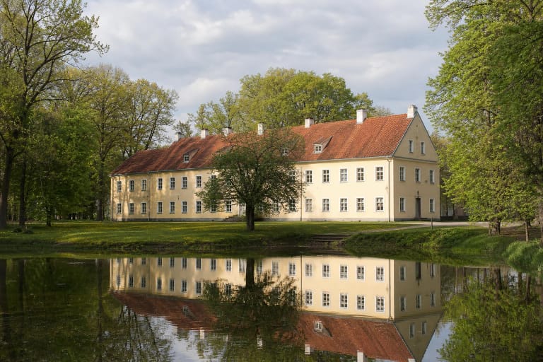 Geltonas Akmenių dvaro buvusios oficinos pastatas su raudonų čerpių stogu atsispindi ramiame tvenkinyje