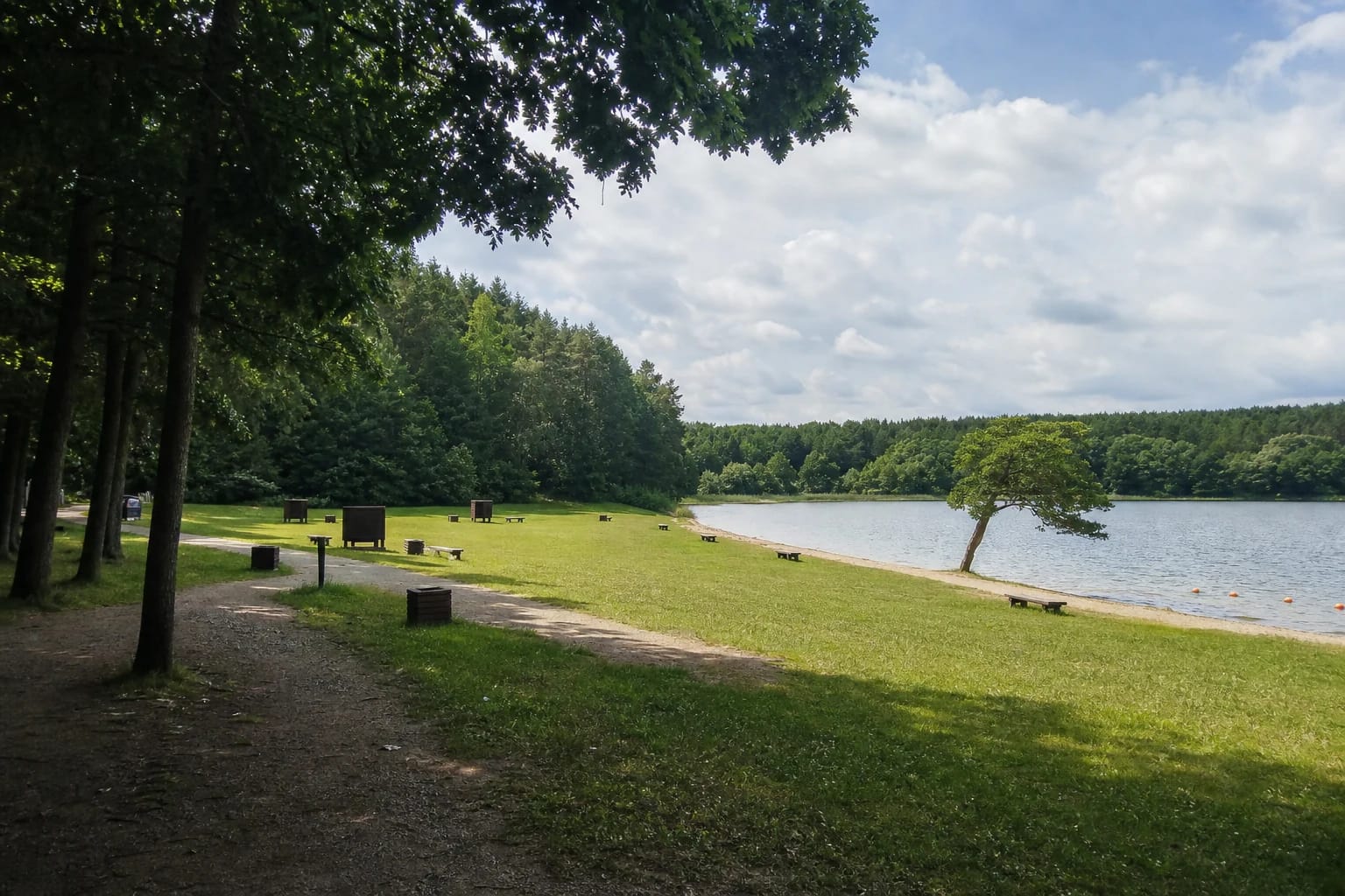 Lake Akmena Beach with a green shore, sandy strip, benches, and a wooded opposite bank