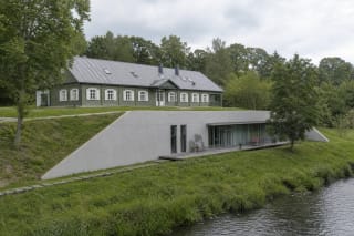 Restored timber Anykščiai Arts Incubator on a green slope, with its glazed lower level opening towards the Anykšta stream