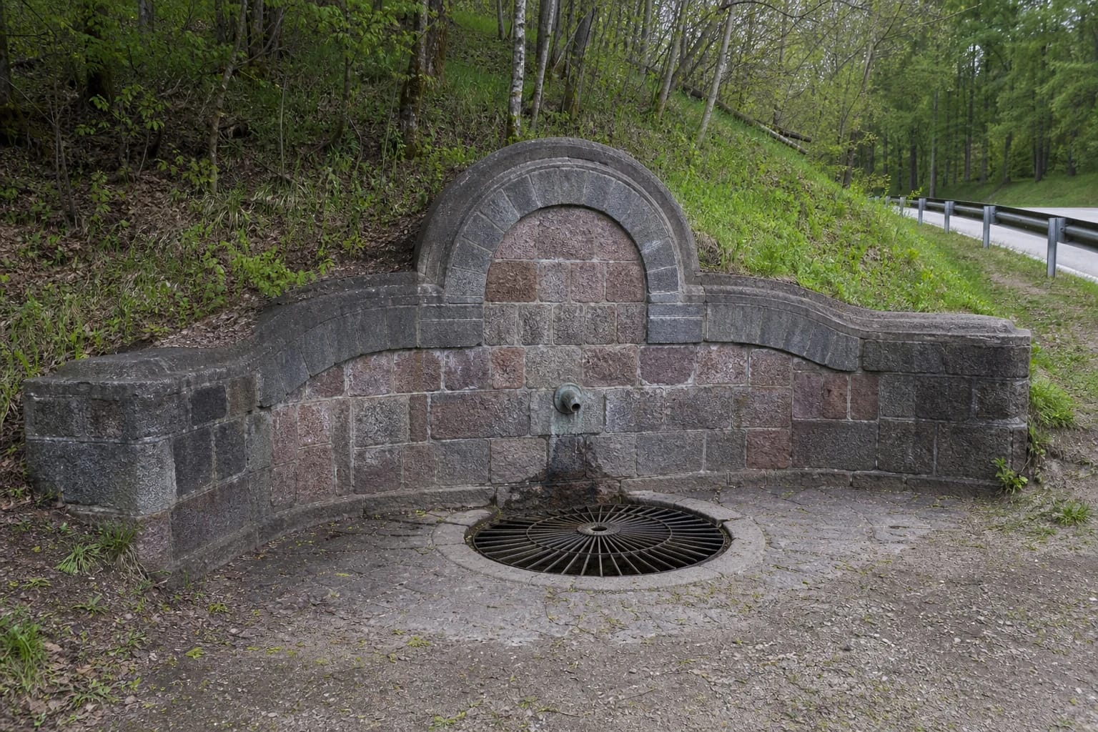Ariogala Spring with a stone wall, dark central stone, steps, and bench on a wooded Dubysa Valley slope