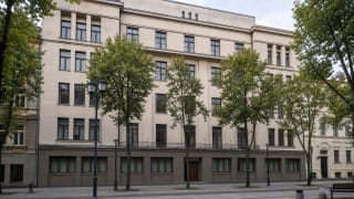 Светлый пятиэтажный фасад здания на аллее Лайсвес, 13 с прямоугольными окнами, неглубокими ризалитами и деревьями