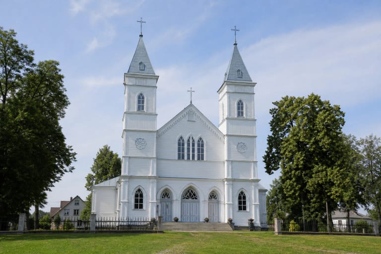 Balta Aukštadvario Kristaus Atsimainymo medinė bažnyčia su dviem bokštais, veja ir medžiais