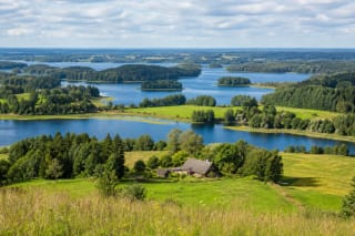 Aukštaitija lake-district panorama with linked blue lakes, forested islands, rolling fields, and a traditional timber homestead