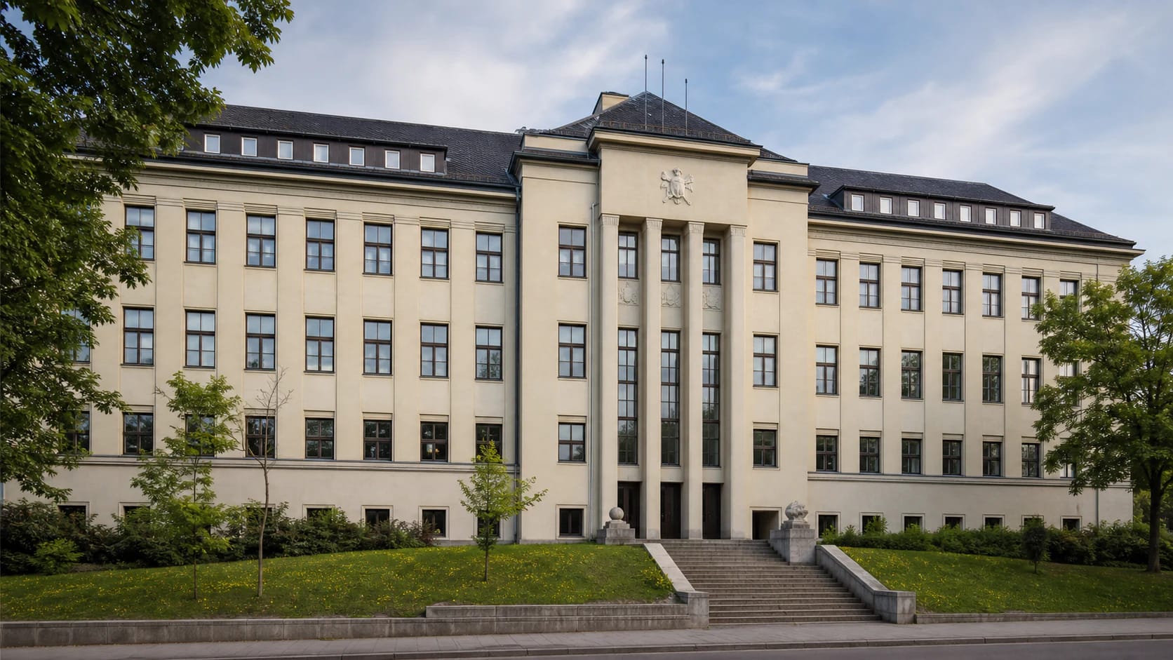 Long light interwar facade of the Higher Technical School in Kaunas with a central projection, stairs, and green Tvirtoves aleja surroundings