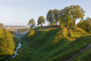 Žalias Bakainių piliakalnio kompleksas su mediniais laiptais, gynybiniais pylimais ir vingiuojančia Liaudės upe