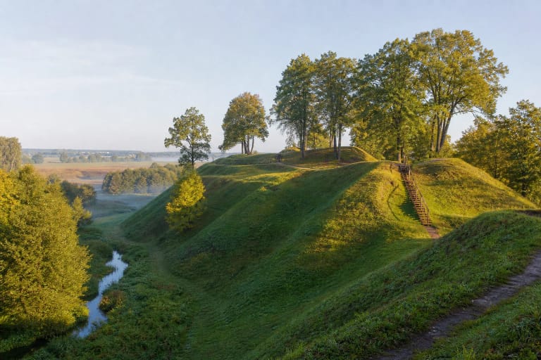 Žalias Bakainių piliakalnio kompleksas su mediniais laiptais, gynybiniais pylimais ir vingiuojančia Liaudės upe