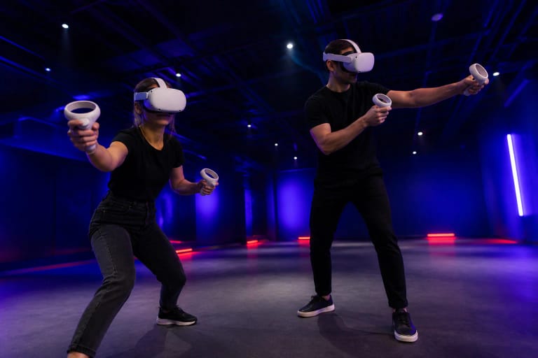 Du žaidėjai su baltais VR akiniais ir valdikliais tamsioje mėlynai violetinėje Bala VR erdvėje