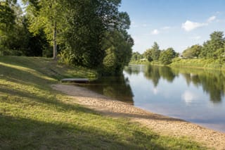 Sandy Berčiūnai Beach beside the calm Sanžilė Canal, surrounded by green trees and a grassy slope