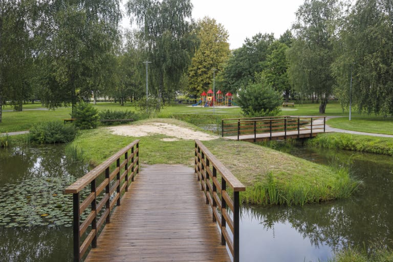 Du mediniai tilteliai per Beržynėlio parko tvenkinį į žalią salą, tolėliau matyti beržai, takai ir raudonstogė vaikų žaidimų aikštelė