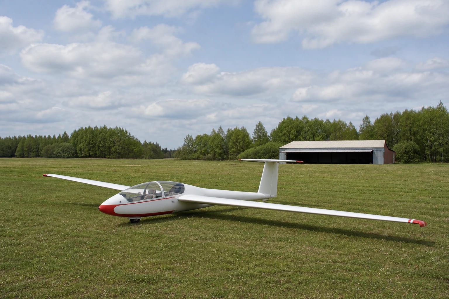 White glider with red trim on Biržai Aeroclub's grass airfield beside a hangar