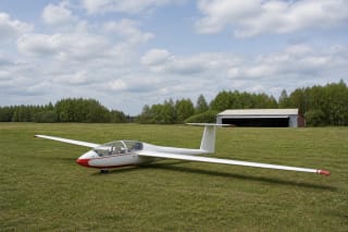 White glider with red trim on Biržai Aeroclub's grass airfield beside a hangar