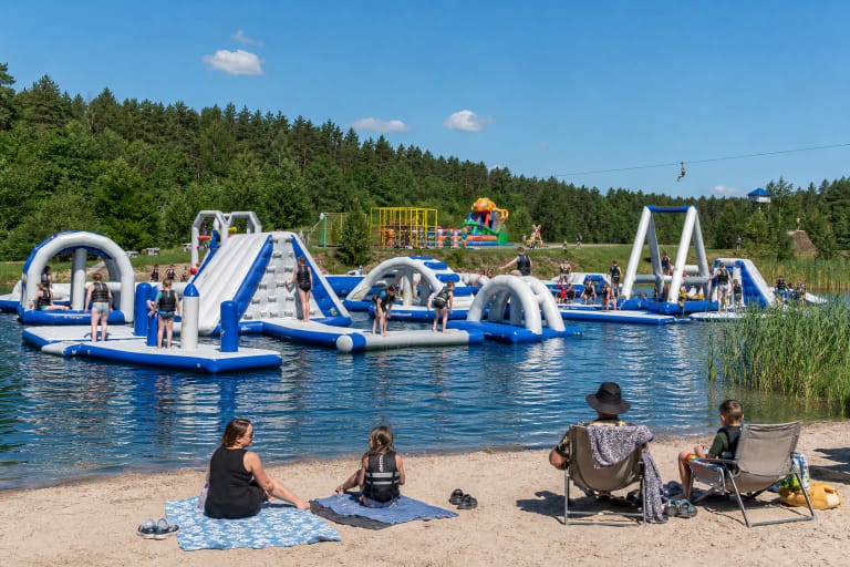Blue-and-white floating obstacle course at BOOM Park in Žibininkai, with families on the sandy shore and forest around the pond