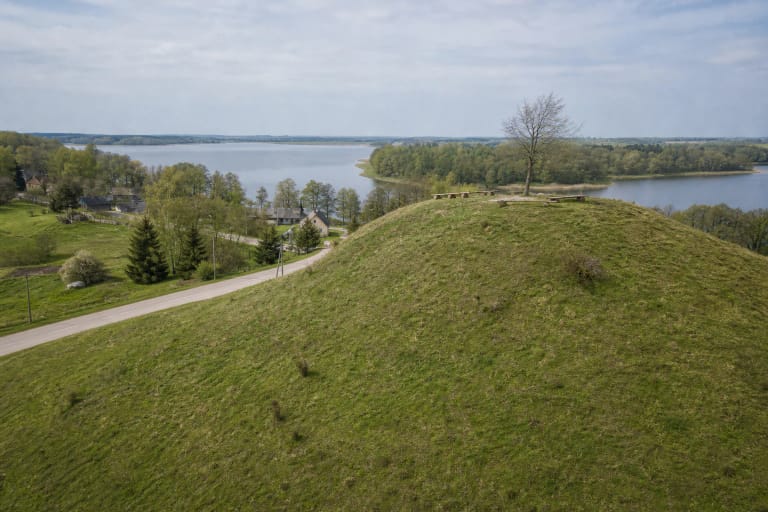 The green slope of Buteliūnai Hillfort with a lone tree above the village road and Lake Prapuntas visible beyond