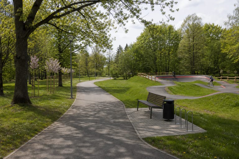 Platus asfaltuotas Dainų parko takas tarp brandžių medžių, jaunos žydinčios sakuros ir juoda pump track trasa su raudonu kraštu