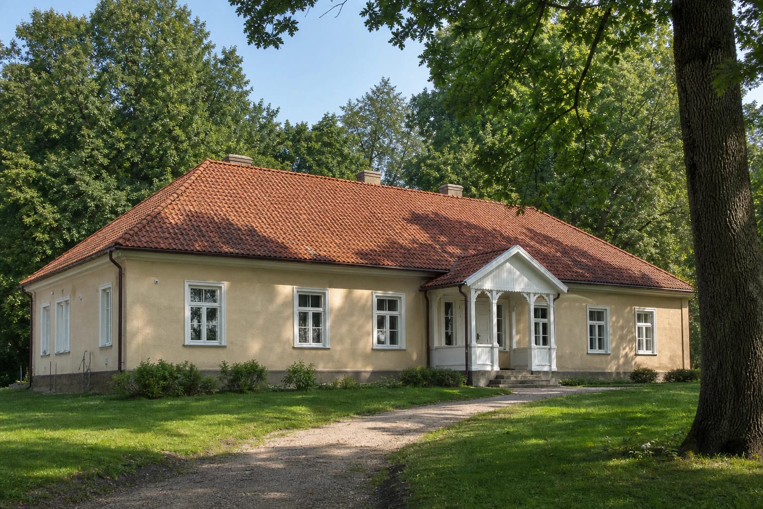 Long pale-plastered Dapkiškės manor house with a red-tile roof and white wooden porch beneath mature trees