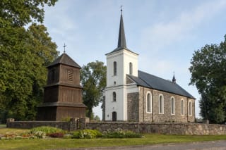 Каменная церковь Дарбеная с высокой белой башней, отдельной трёхъярусной деревянной колокольней и оградой церковного двора из полевых камней