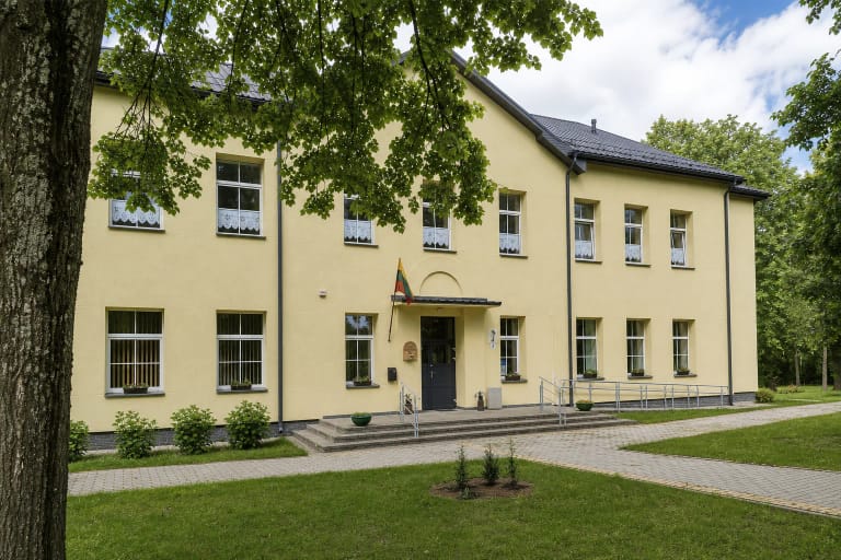 Pale-yellow Dargužiai Crafts Centre beneath leafy trees, with a Lithuanian flag and an accessible entrance ramp