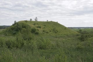 Open grassy southeastern slope of Dirvonai Hillfort above a brush-filled wet hollow among the fields of the Sudovian Upland