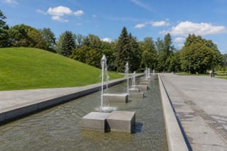 The shallow fountain basin in Draugystė Park, Kaunas, with vertical water jets, concrete blocks, a grassy mound, and a broad paved route