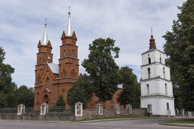 Dusetos Holy Trinity Church in red brick with two tall towers and a separate white eighteenth-century bell tower among trees