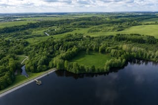 Aerial view of Eketė Hillfort's large green enclosure, tree-covered rampart, and reservoir wrapping around three sides
