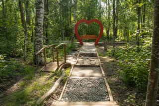 Akmenukų, smėlio ir medinių ritinių atkarpomis per žalią Drąseikių mišką vedantis Eko basų kojų parko takas su raudona širdies arka