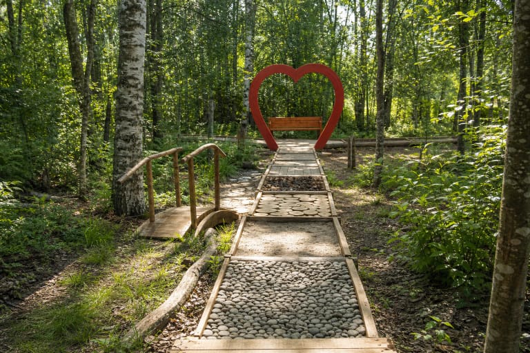 Akmenukų, smėlio ir medinių ritinių atkarpomis per žalią Drąseikių mišką vedantis Eko basų kojų parko takas su raudona širdies arka