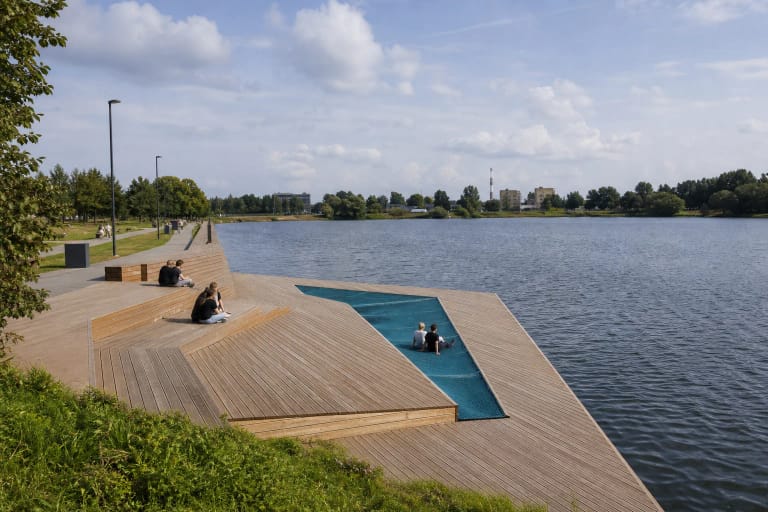 Timber terrace on the Ekranas Reservoir shore with a blue net lounging strip above calm water in Panevėžys