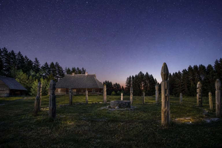 Aukštaitiška medinė sodyba ir apskrita dangaus šviesulių stebykla su mediniais ženklais vakaro danguje