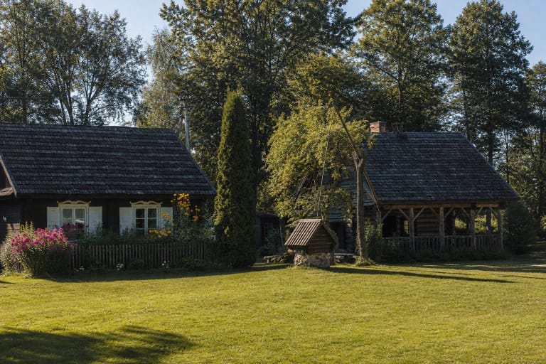 Etnografinės Suvalkiečio sodybos tamsių rąstų namas, balti langai, medinis kluonas ir šulinys erdviame žaliame kieme