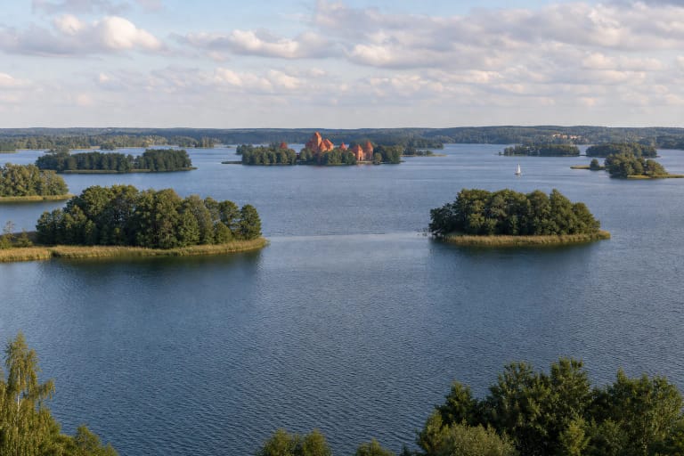 Galvės ežeras su miškingomis salomis, ramiu vandeniu ir tolumoje matoma Trakų salos pilimi