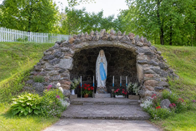 Gardamo Lurdo lauko akmenų grota su balta ir mėlyna Marijos Lurdiškės skulptūra, žvakėmis ir gėlėmis žaliame šlaite