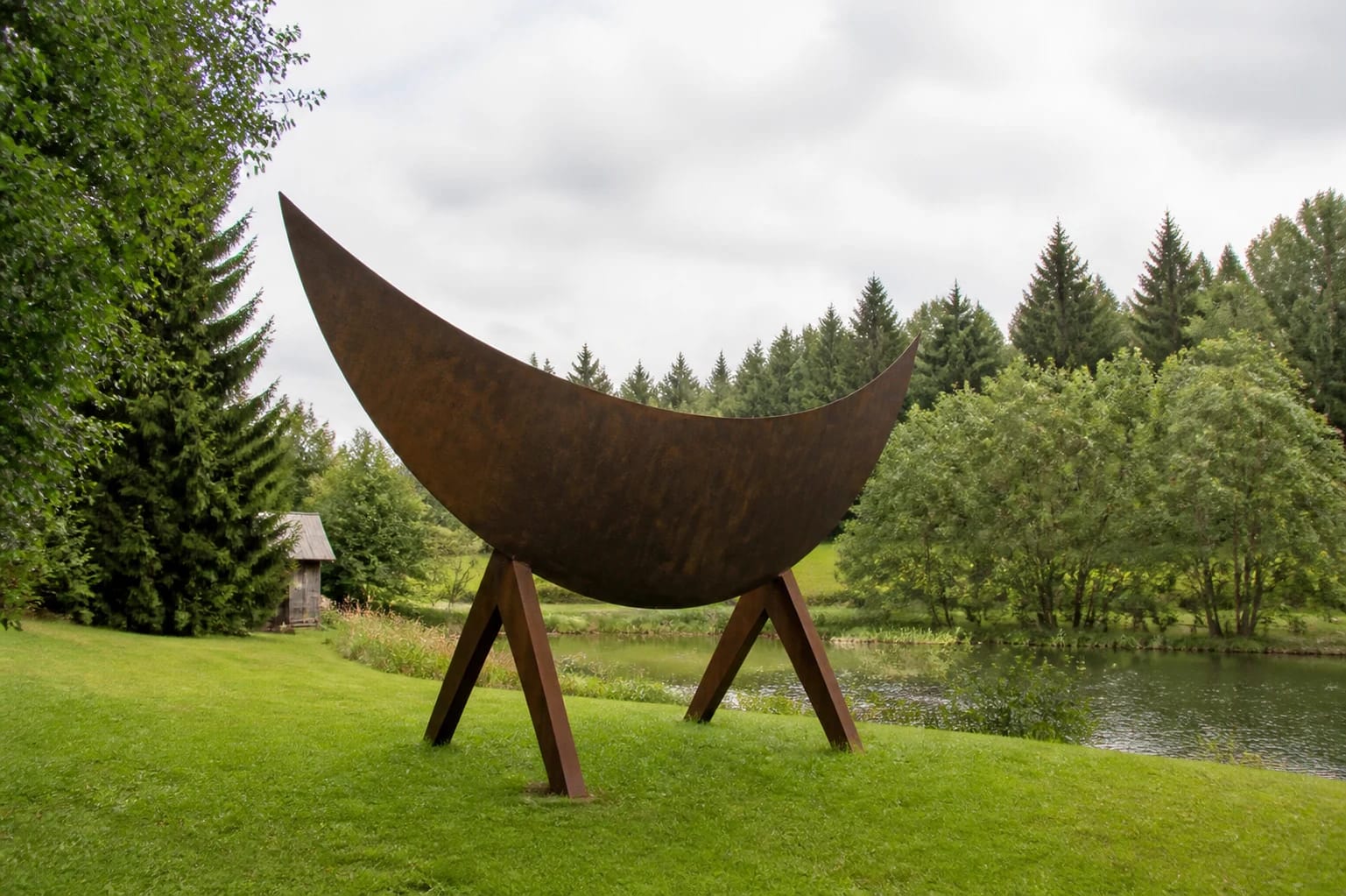 Giedrius Mazūras Sculpture Park's weathered steel sculpture on three supports beside a pond and green lawn in Antalgė
