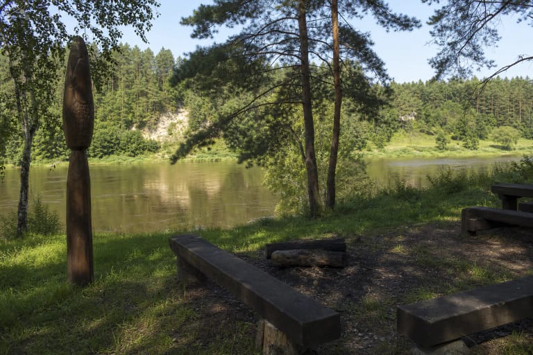 Giraitiškių poilsiavietės mediniai suolai ir žuvies skulptūra pušų pavėsyje prie Nemuno, kitapus matant Škėvonių atodangą