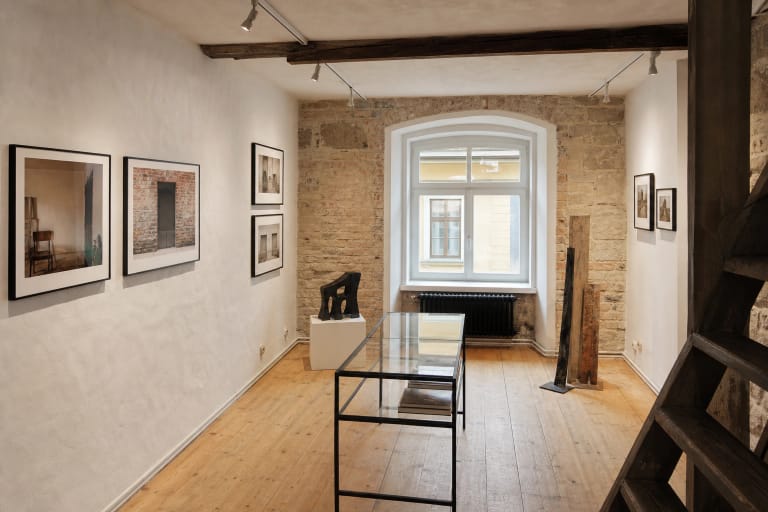 Kompaktiška GODÒ galerijos salė su fotografijos darbais, senų plytų siena ir kuklia šiuolaikine skulptūra