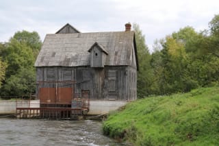 Grey vertical-board walls, broad gabled roof, brick chimney, and hydroelectric water channel at Gudai Water Mill beside the Virvytė