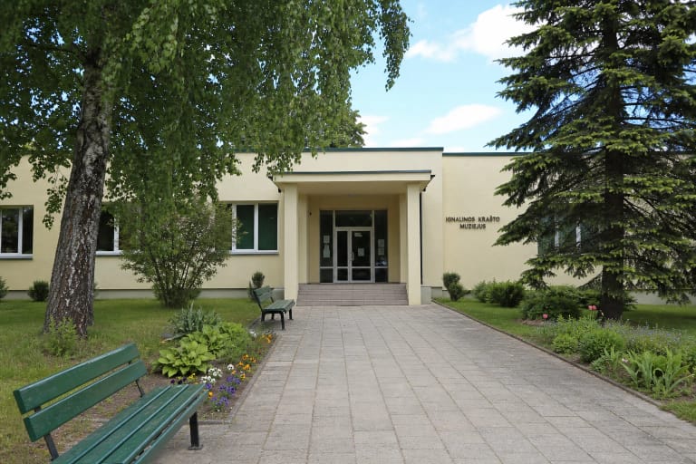 Šviesiai gelsvas Ignalinos krašto muziejaus fasadas, stiklinis įėjimas, grįstas takas ir žalios suoliukų eilės tarp medžių
