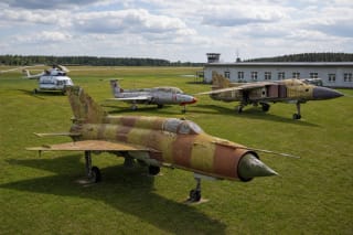Įstros lėktuvų muziejaus lauko ekspoziciją vaizduojantis kadras su aptrintu kamufliažiniu MiG-21, L-29, MiG-23 ir Mi-8 aerodromo pievoje