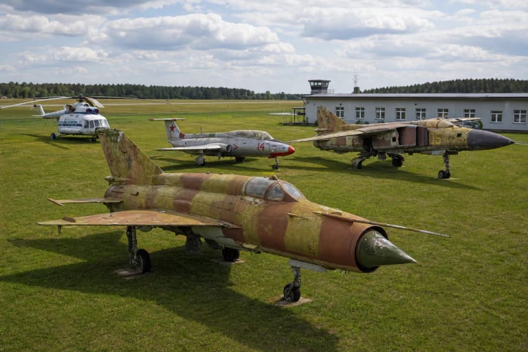 Įstros lėktuvų muziejaus lauko ekspoziciją vaizduojantis kadras su aptrintu kamufliažiniu MiG-21, L-29, MiG-23 ir Mi-8 aerodromo pievoje