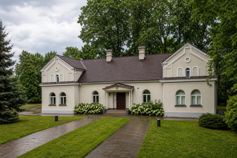 Pilkšvai smėlinis simetriškas Janinos Monkutės-Marks muziejaus-galerijos fasadas, tamsus čerpių stogas ir žydinčios hortenzijos parke