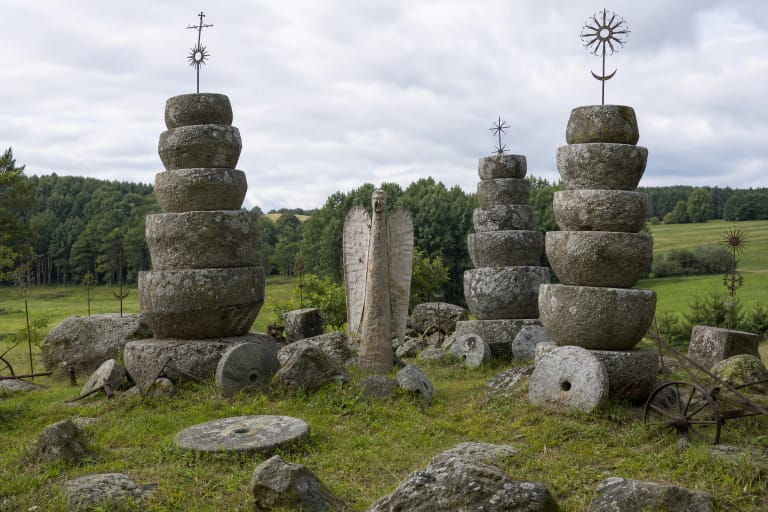 Jotvingių kiemo akmenų muziejaus girnų bokštai ir ąžuolinis Angelas žaliame Kybartų kaimo pasienio kraštovaizdyje