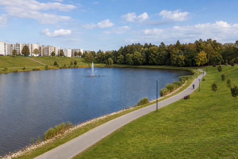 Stadiono Reservoir in Juodpelkis Park among grassy slopes, a curving path, reeds, and apartment buildings in Mažeikiai